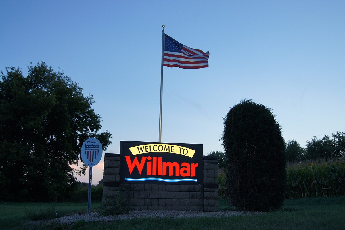 A lit Welcome to Willmar sign on a block base at dusk, with an American flag on a pole above it, shrubs and trees on either side and a cornfield behind