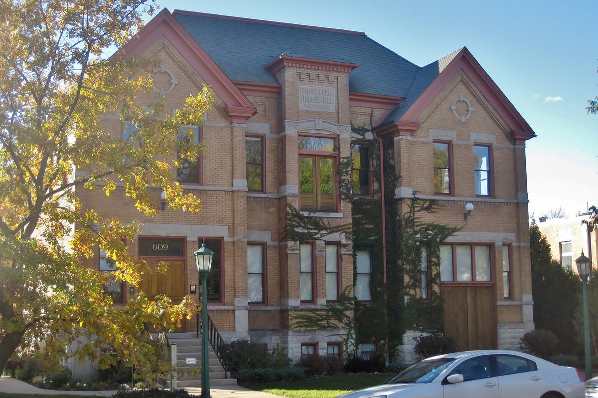 The tan brick Gross Point Village Hall in Wilmette, with red-trimmed gables and ivy on the front, a white car at the curb and autumn leaves on the trees