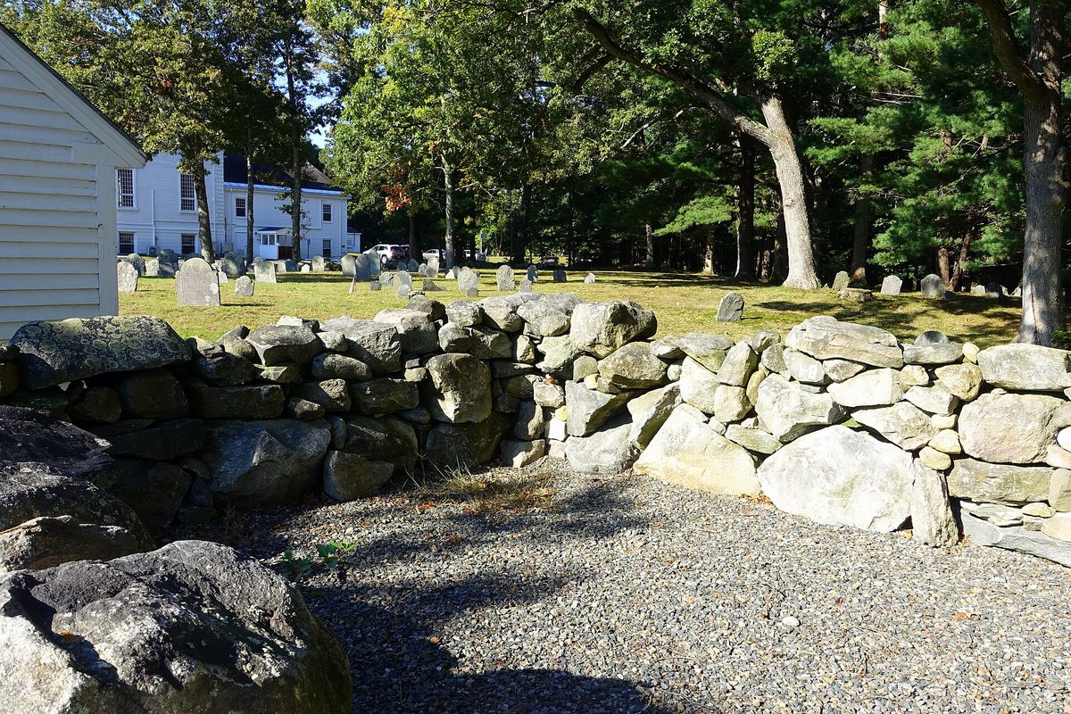 The fieldstone walls of Wilmington's 1814 town pound, with old gravestones on the grass beyond and white houses behind the trees