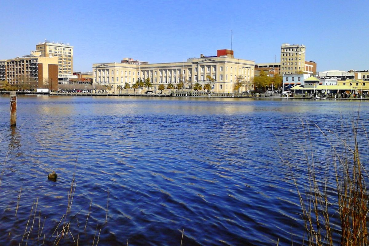 Downtown Wilmington's waterfront seen across the blue Cape Fear River, with a long columned building and taller buildings behind it under a clear sky
