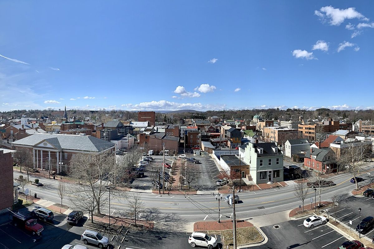 A wide view over downtown Winchester from a parking deck, with brick buildings, a columned building and parking lots below and low hills on the horizon under a blue sky