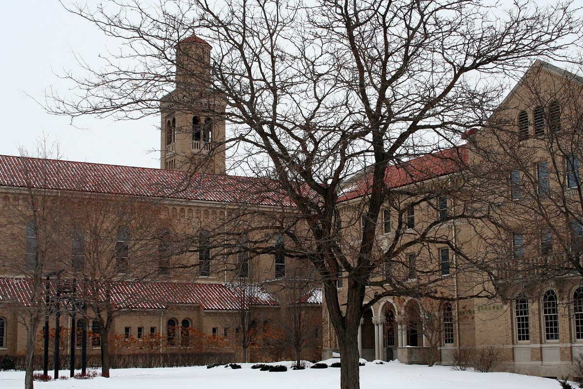 Tan brick buildings with red tile roofs and a square bell tower on the former College of Saint Teresa campus in Winona, behind a bare tree in winter snow