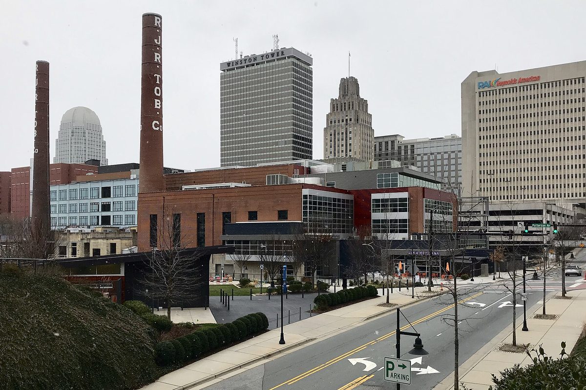 Downtown Winston-Salem skyline behind the old brick Bailey Power Plant and its R.J.R. Tob. Co. smokestacks, with an empty street and a parking sign in front