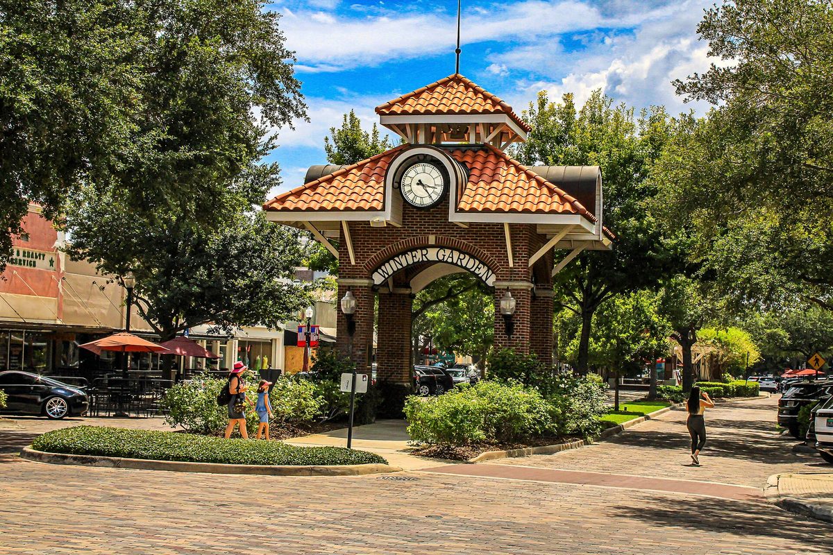 The brick clock tower with a red tile roof and a Winter Garden arch on Plant Street, with shade trees, outdoor umbrellas and people walking by