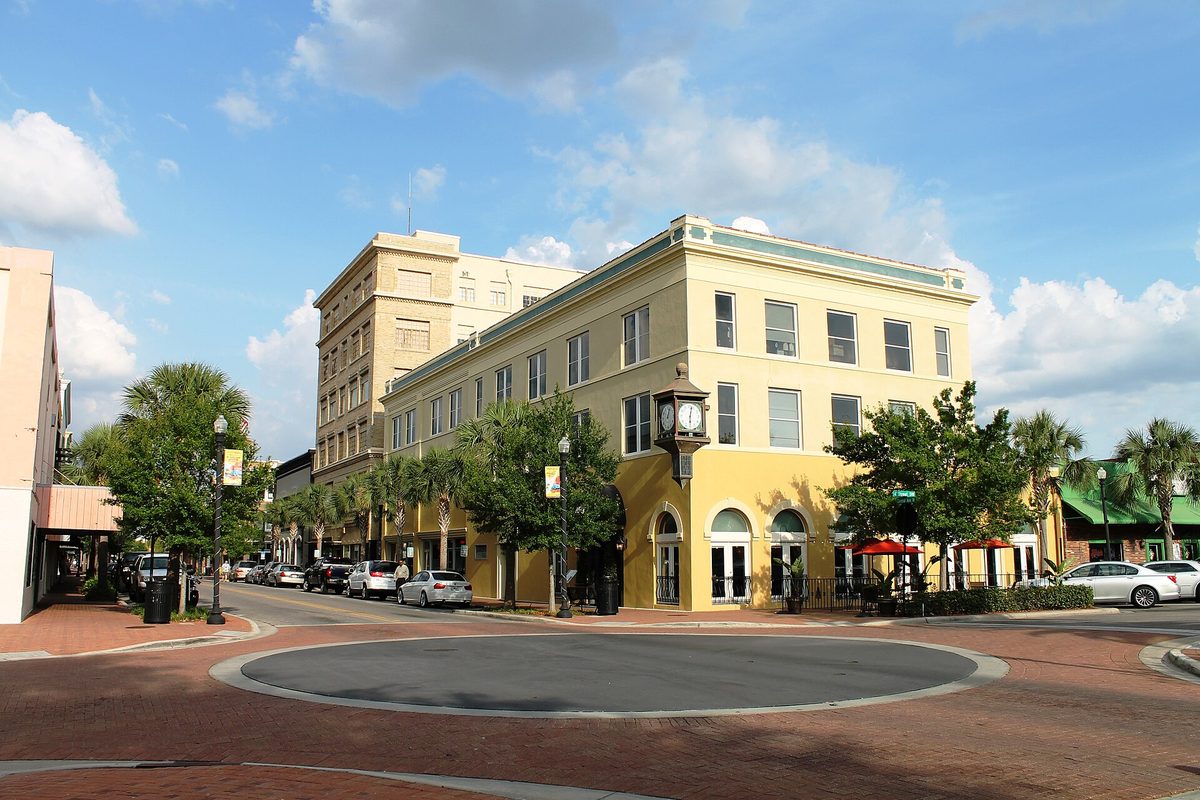 A cream four-story building with a street clock on a post at a brick roundabout in downtown Winter Haven, with palm trees and parked cars under a blue sky