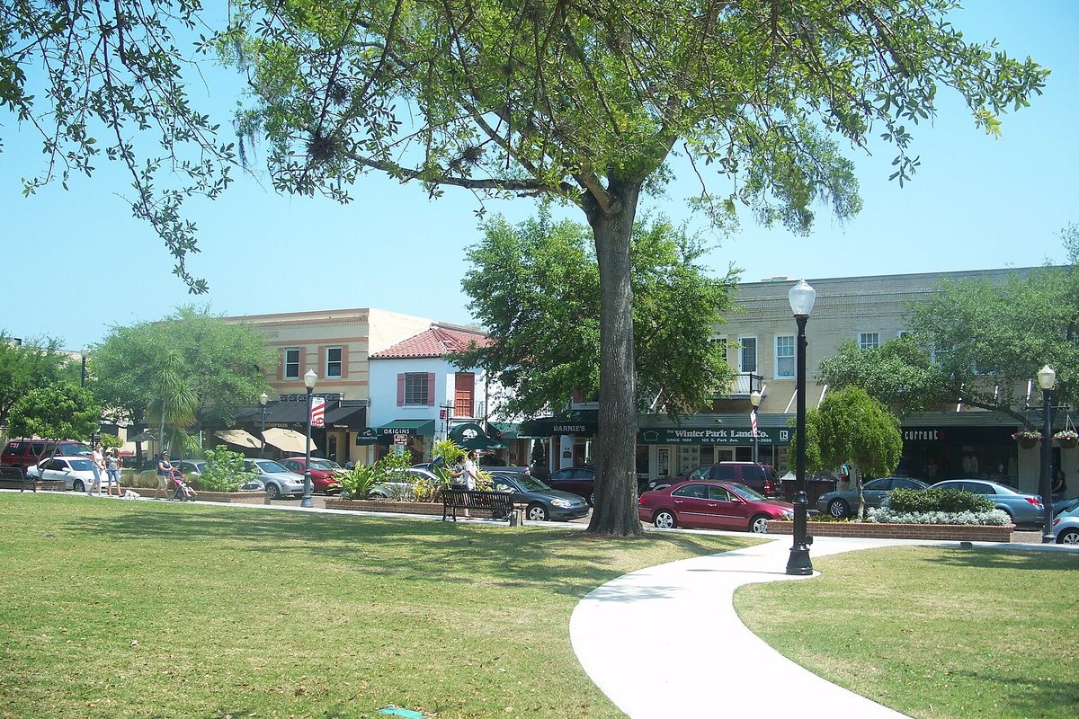 A lawn and curving walkway under a large shade tree in downtown Winter Park, with two-story shops and parked cars along the street behind