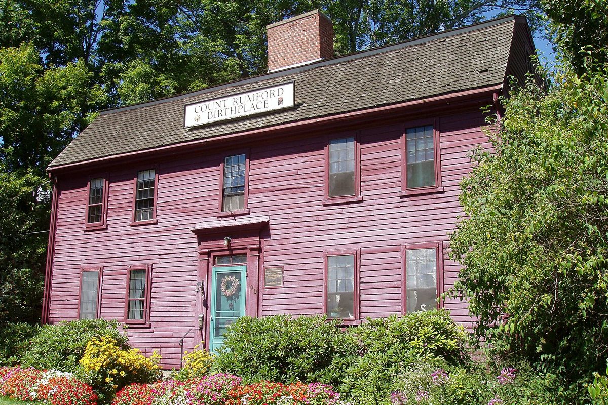 A red clapboard colonial house in Woburn with a sign reading Count Rumford Birthplace, a teal front door with a wreath and bright flower beds