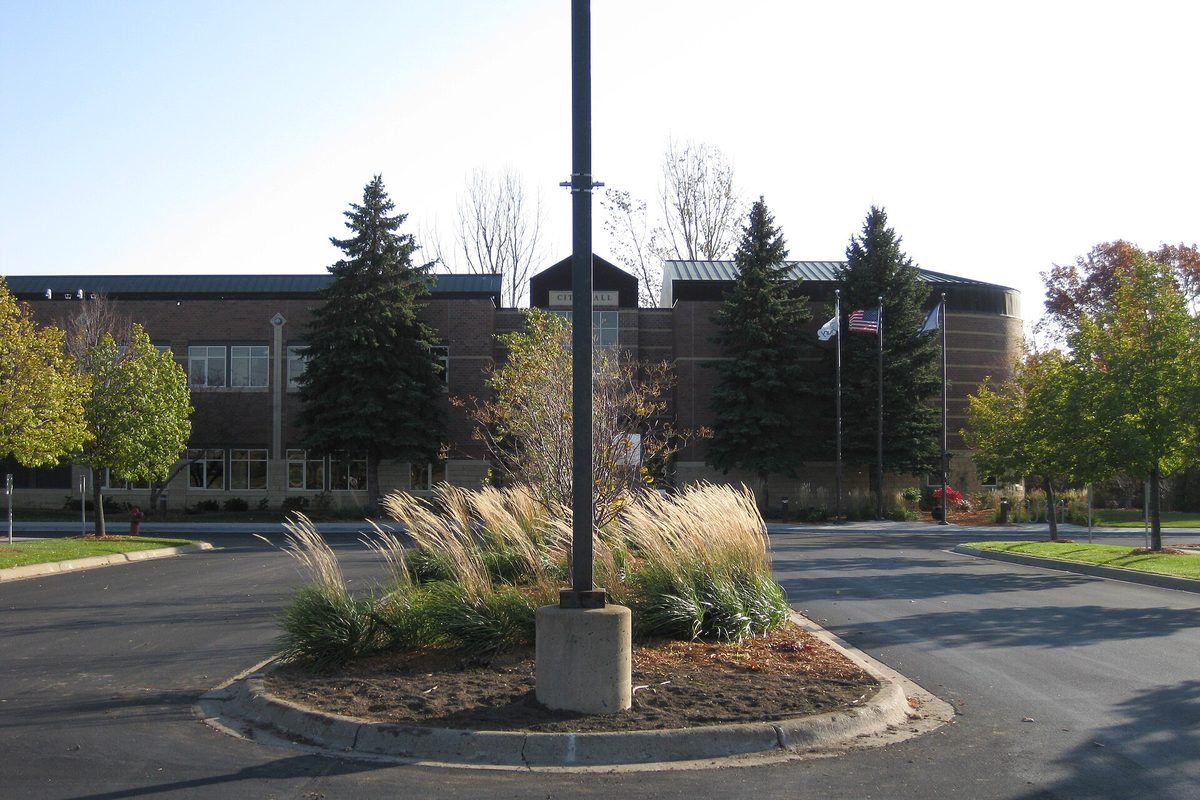 Woodbury City Hall, a brick building with a green roof and flagpoles, behind evergreen trees and a planted island of ornamental grasses