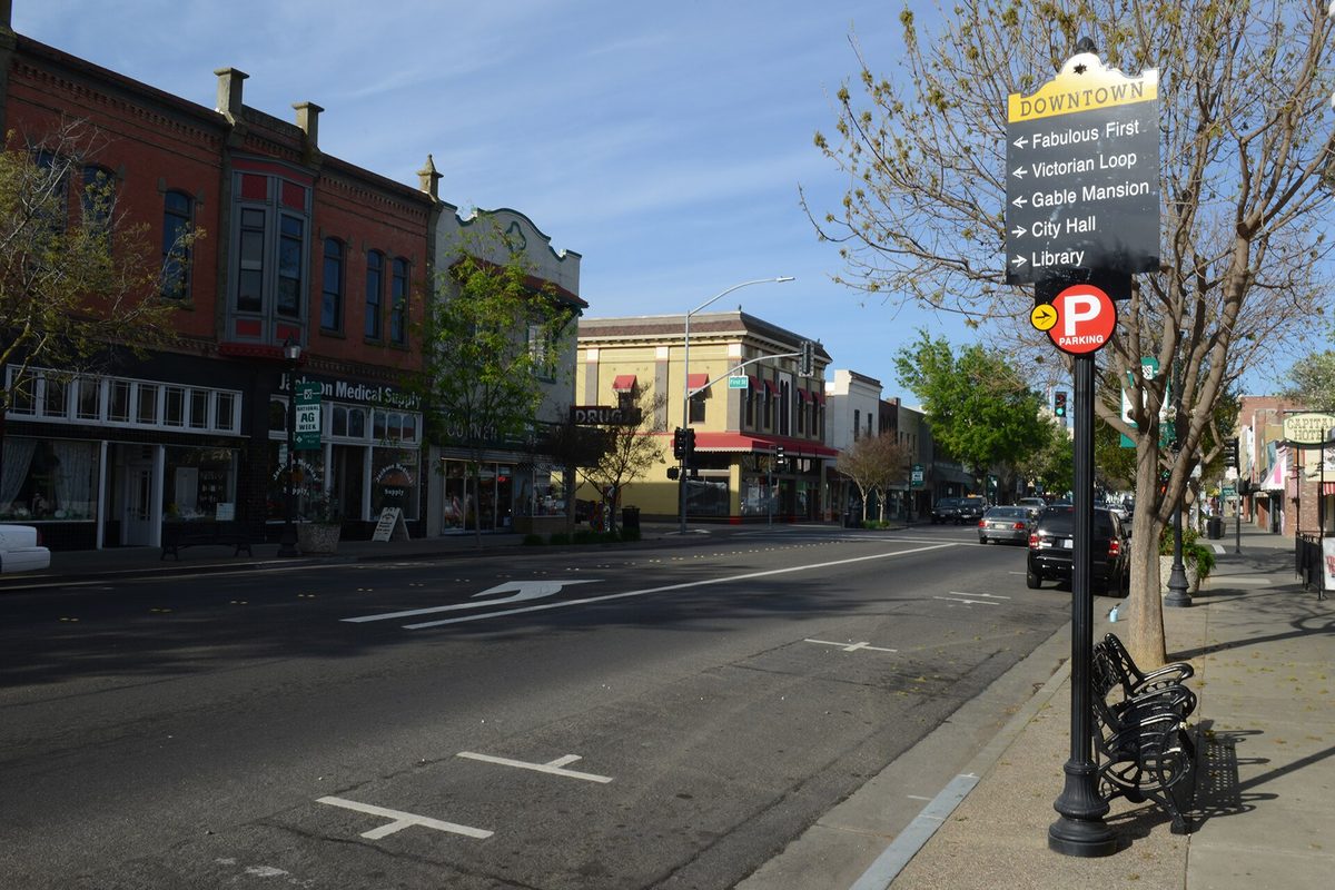 Main Street in downtown Woodland, with brick storefronts, a parking sign and a signpost pointing to the Victorian Loop, Gable Mansion, City Hall and the library