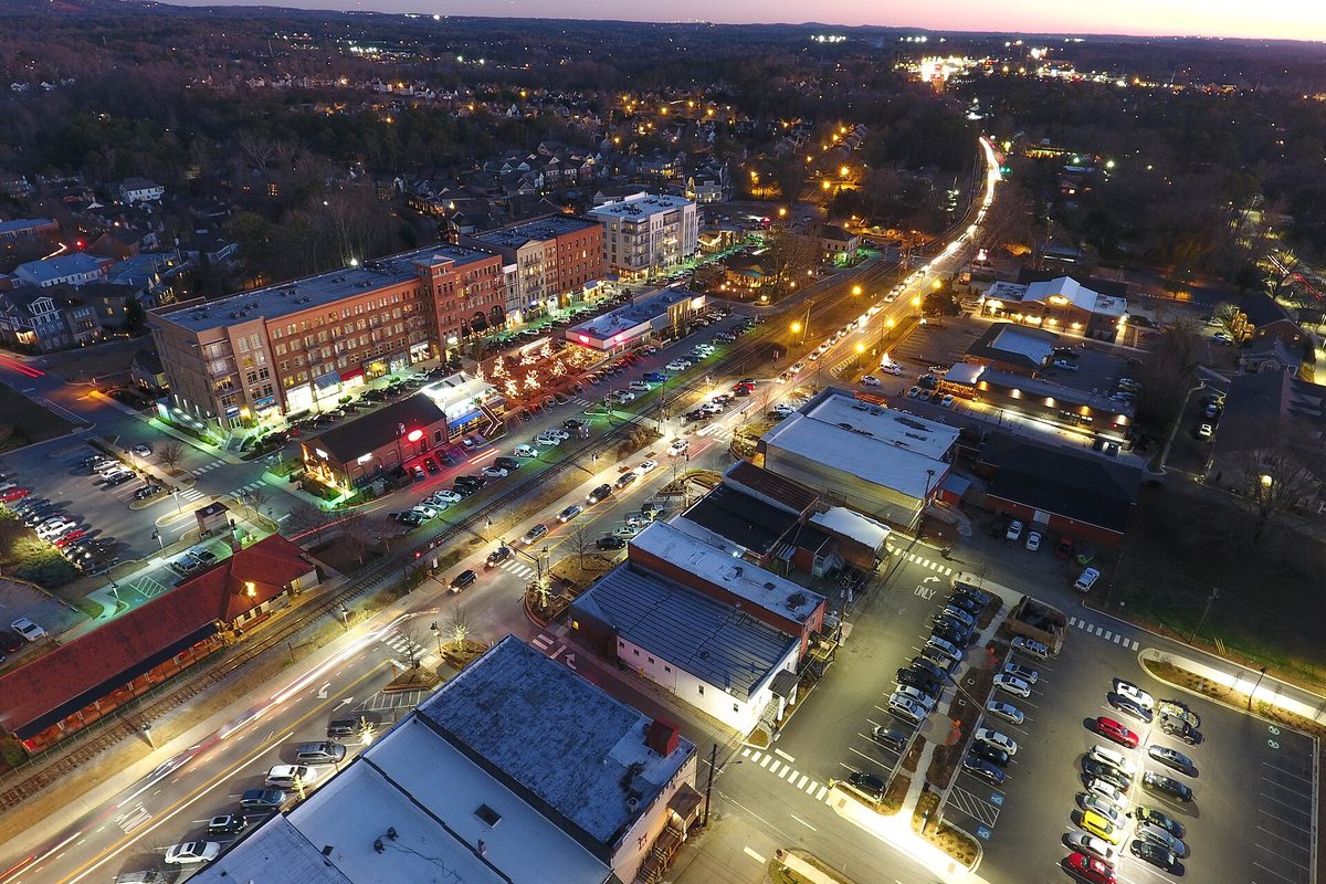 Downtown Woodstock from above at dusk, with lit shops, brick apartment buildings, full parking lots and a rail line beside the main road