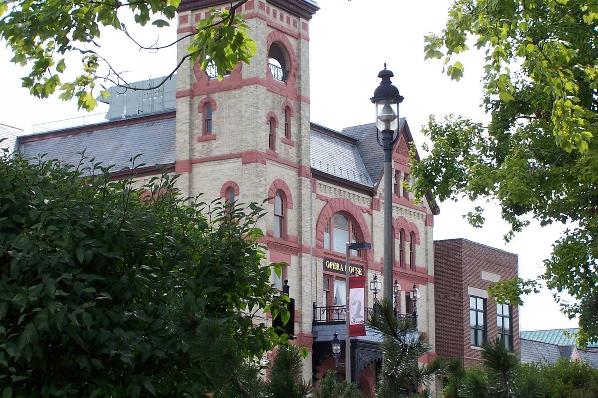 The tower of the tan brick Woodstock Opera House with red-trimmed arched windows, seen through green trees beside an old-style street lamp