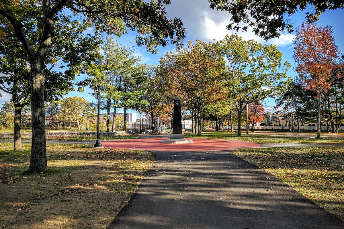 A paved path through Woonsocket's World War II Veterans Memorial Park leading to a dark stone monument on a red plaza, under trees turning orange and red