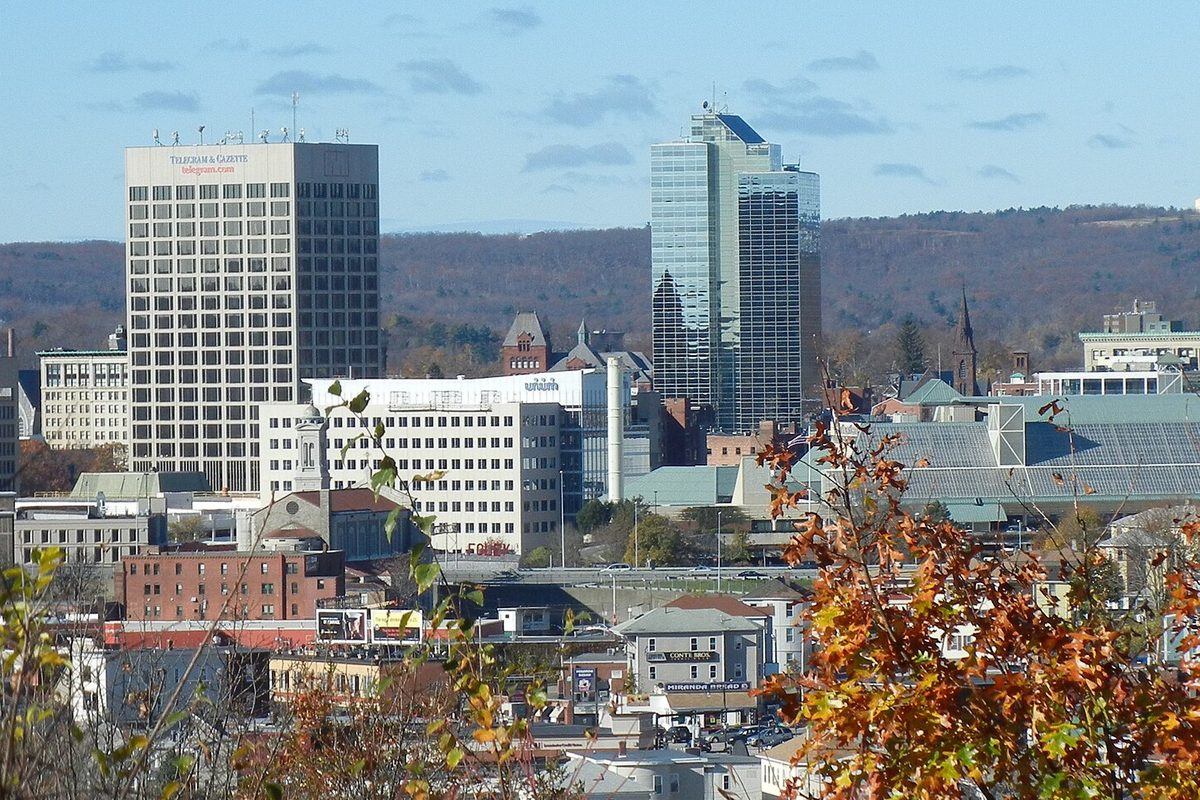 Downtown Worcester seen from a hillside, with two office towers above older brick buildings, autumn leaves in front and wooded hills behind