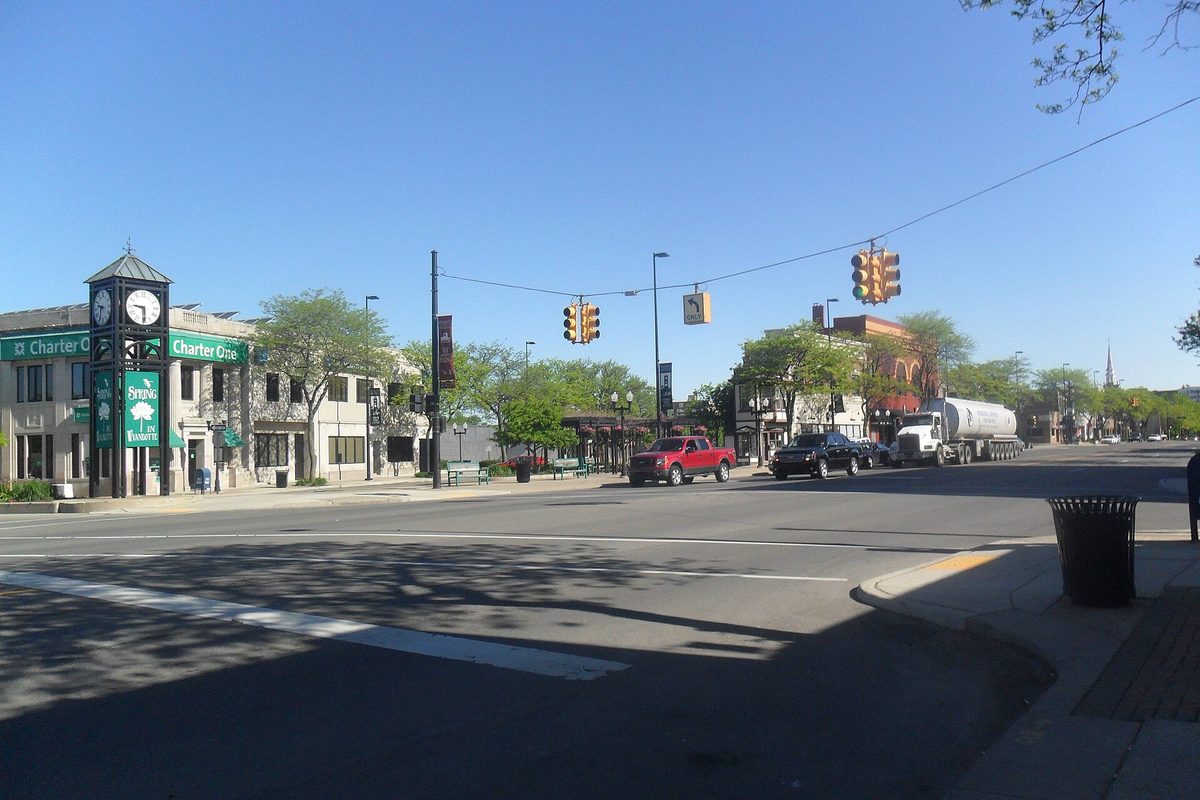 Downtown Wyandotte street on a sunny day, with a green clock tower outside a corner bank, storefronts, traffic lights and a red pickup truck in traffic