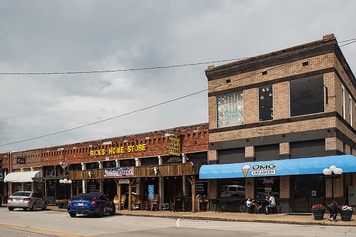 Brick storefronts along downtown Wylie's main street, with a two-story corner building under a blue awning and cars parked at the curb beneath a gray sky