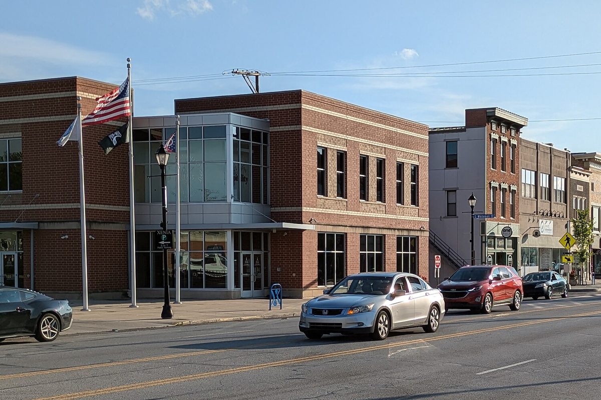 The brick and glass Xenia City Administration Building with flags out front, beside older brick storefronts on East Main Street with cars passing
