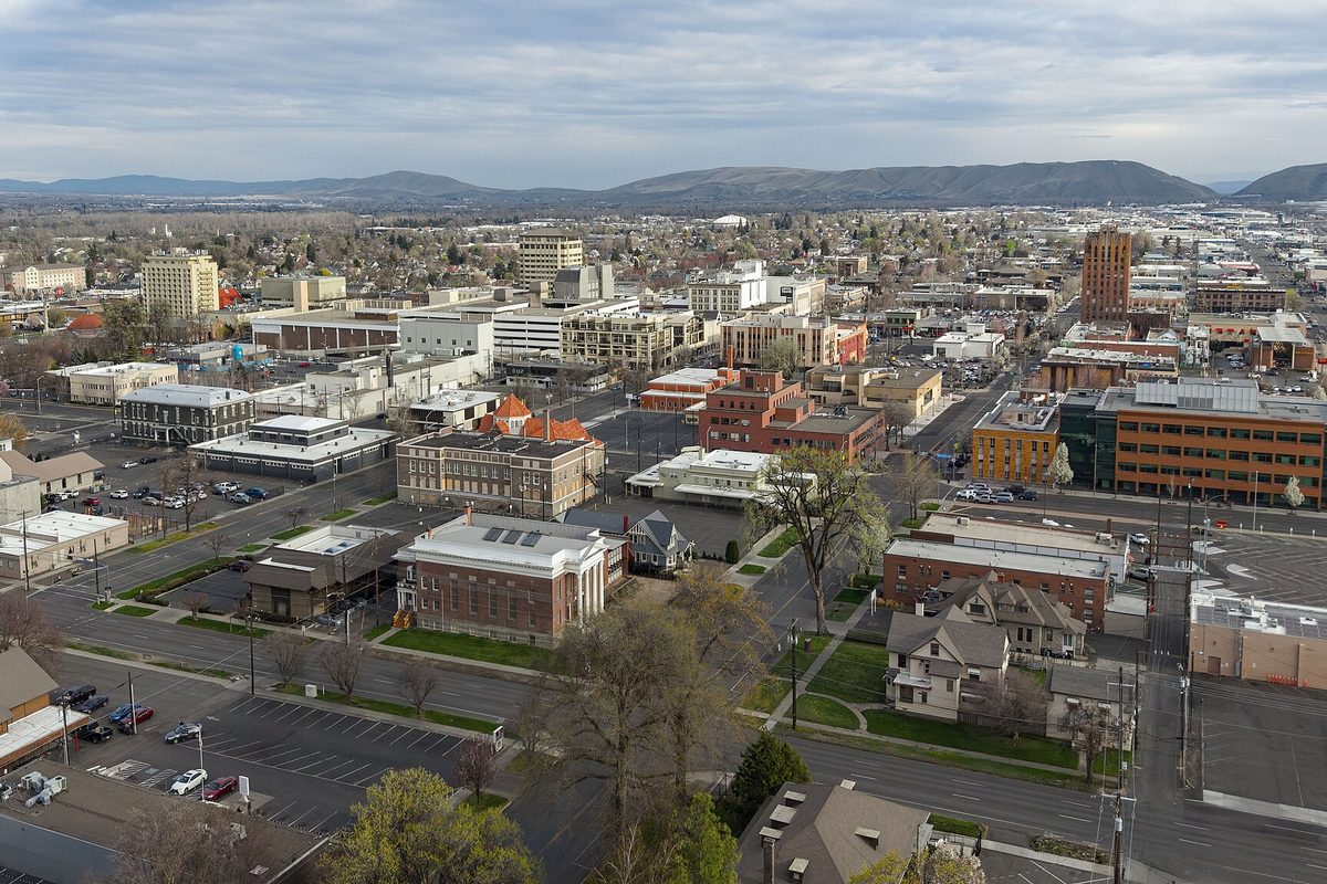 An aerial view over downtown Yakima's brick buildings, parking lots and tree-lined streets, with brown hills on the horizon under a cloudy sky