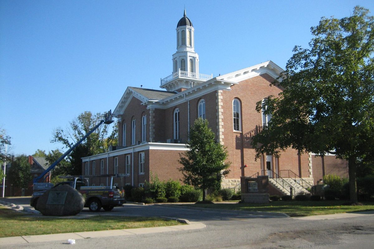 The red brick Kendall County Courthouse in Yorkville with its white cupola, a bucket truck parked beside it and trees under a clear blue sky