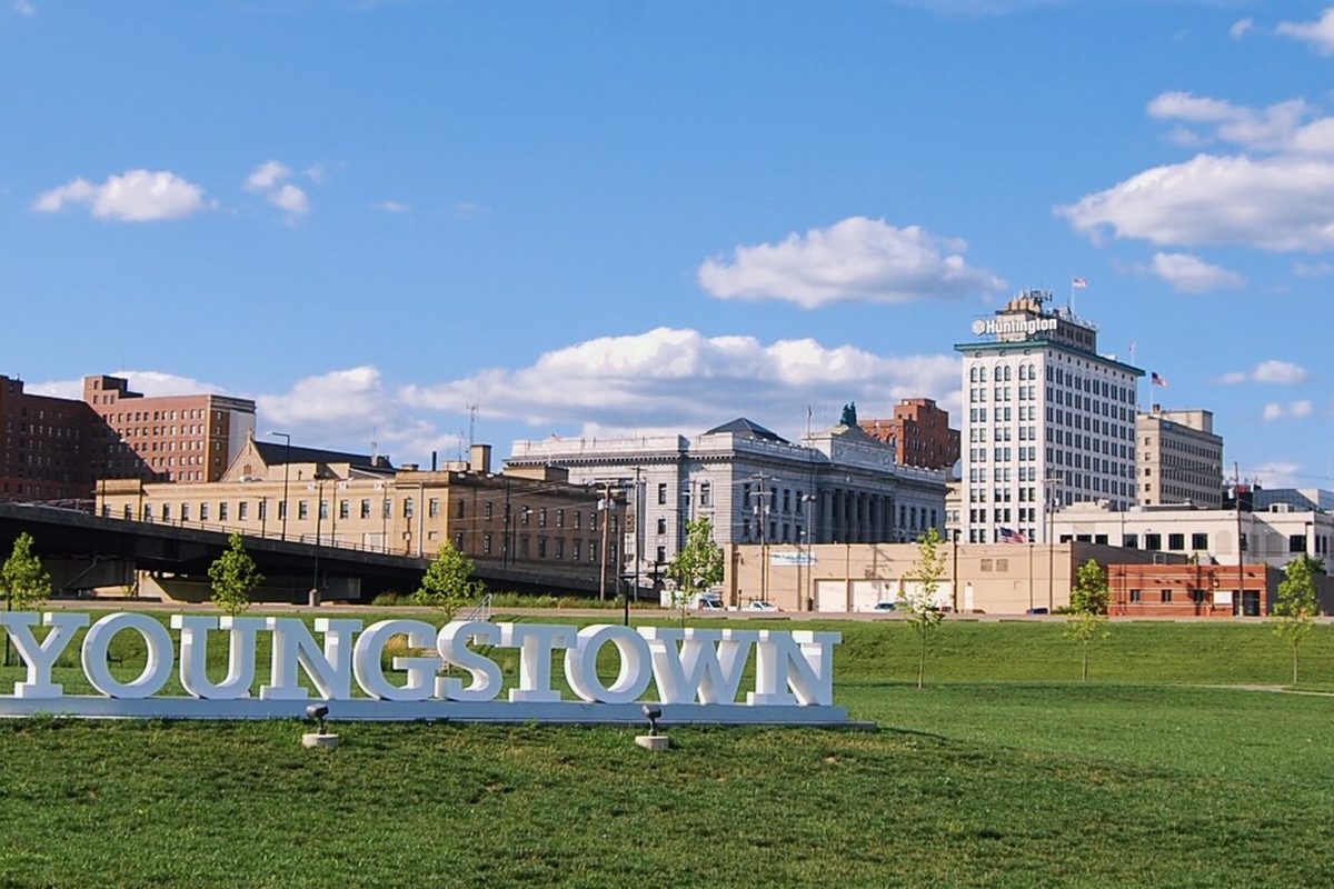 Large white YOUNGSTOWN letters on the lawn of Wean Park, with downtown's brick and stone buildings and a white office tower behind them