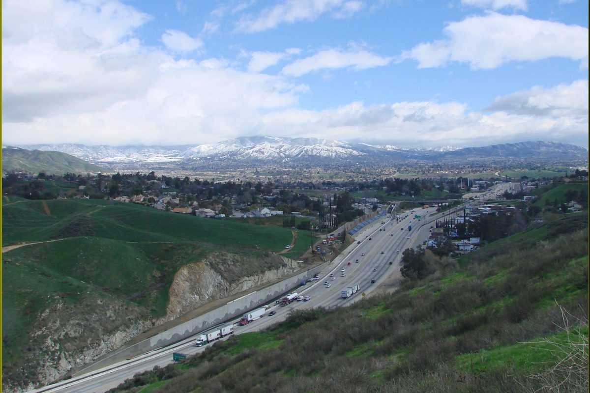 Interstate 10 climbing the Yucaipa Grade through green hills, with snow on the mountains beyond and trucks in the eastbound lanes