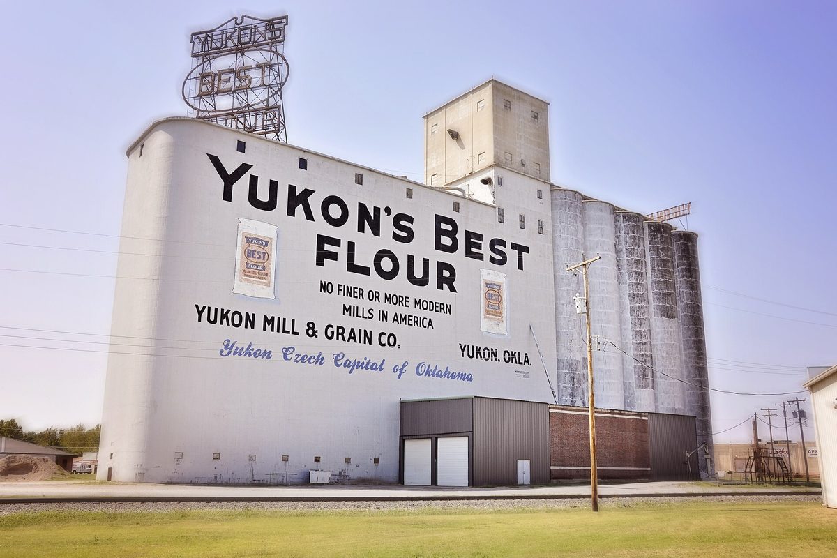 The white Yukon's Best Flour mill and grain elevator, painted with Yukon Mill and Grain Co. lettering, beside a railroad track under a hazy sky
