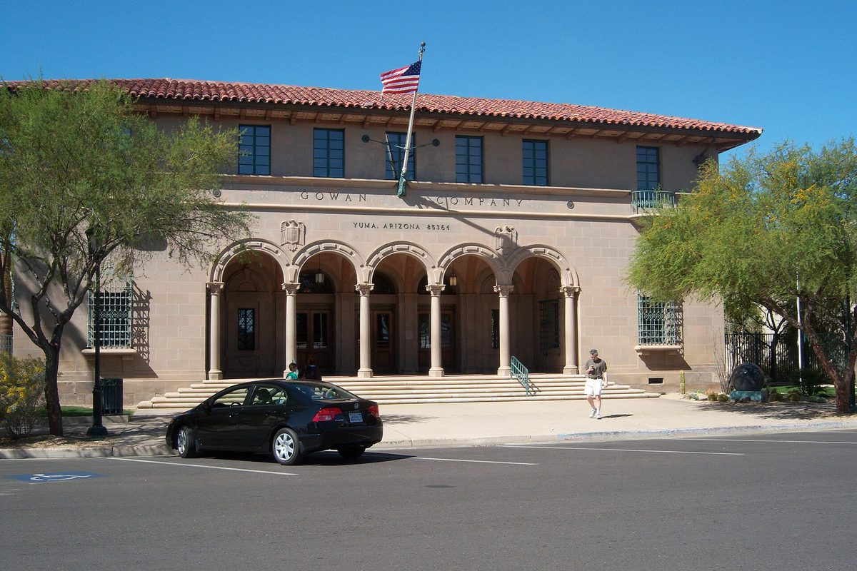 The historic Gowan Company Building in Yuma, a tan former post office with arched entrances and a red tile roof, a U.S. flag on top and a black sedan parked in front