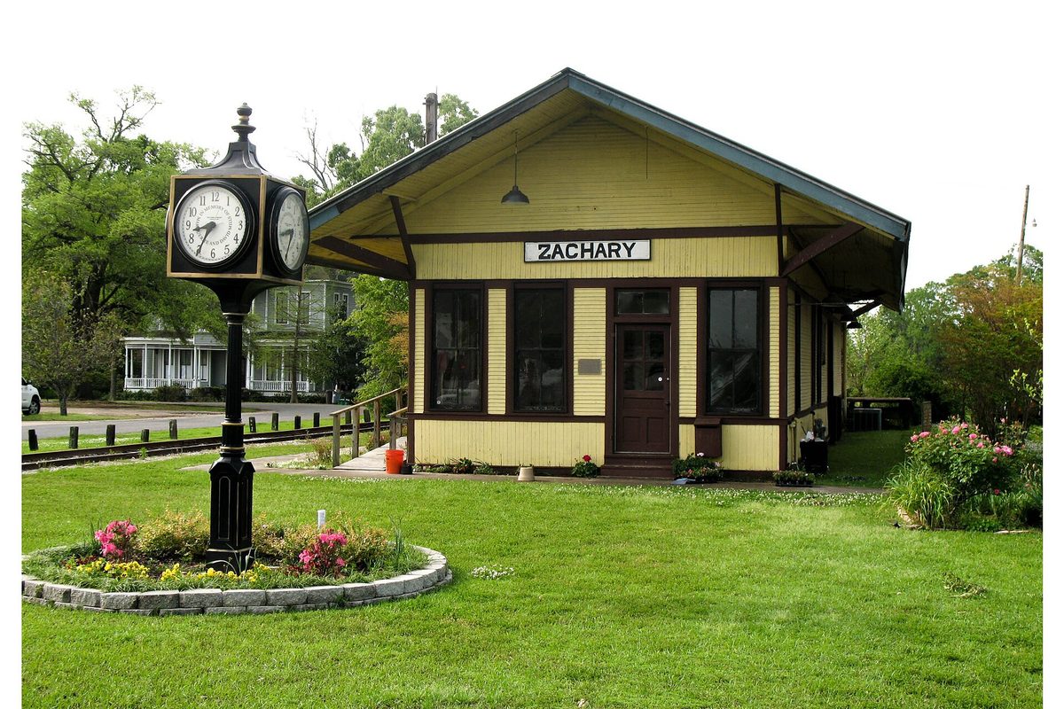 The yellow Zachary railroad depot with a black street clock and a round flower bed on the lawn in front