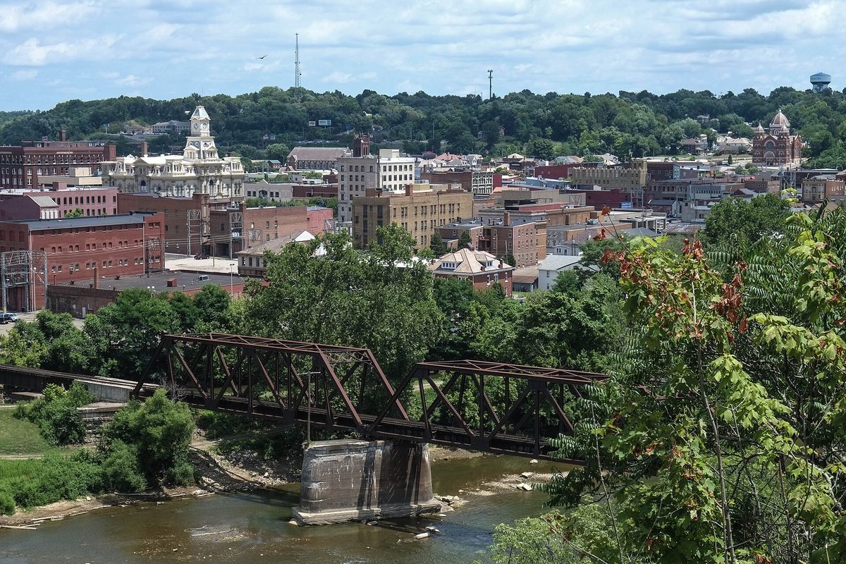 Downtown Zanesville seen from a wooded hillside, with a rusted steel truss bridge on a stone pier crossing the river in front and a domed building on the skyline