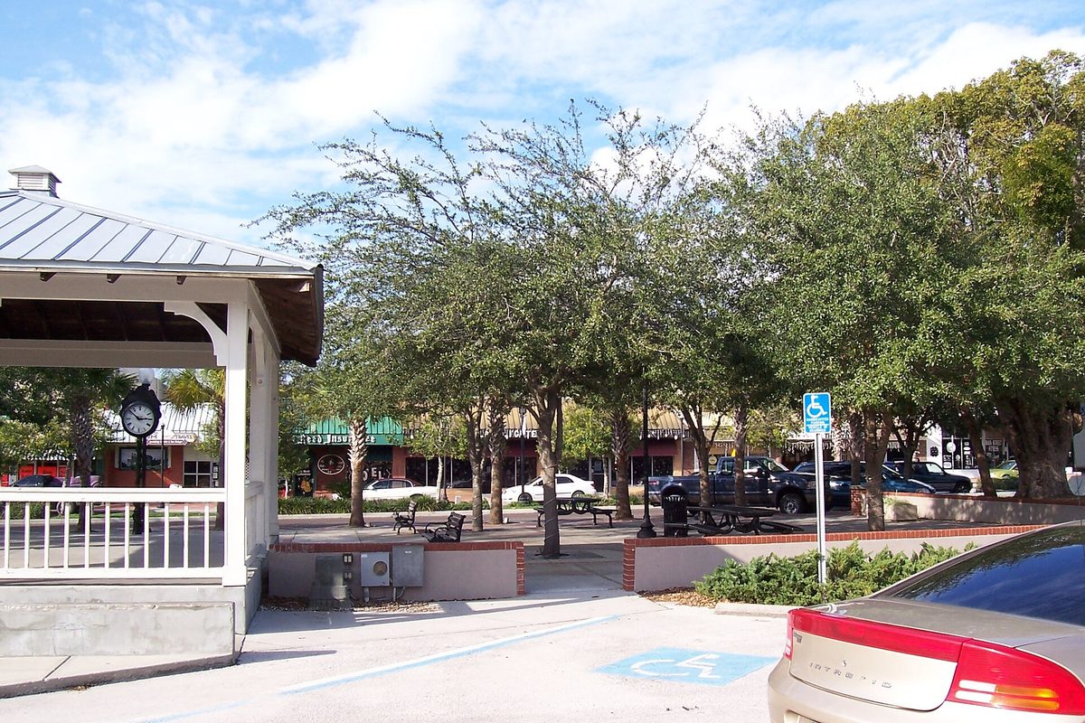 A white gazebo with a street clock on 5th Avenue in downtown Zephyrhills, with shade trees, benches, brick planters and parked cars along the shops