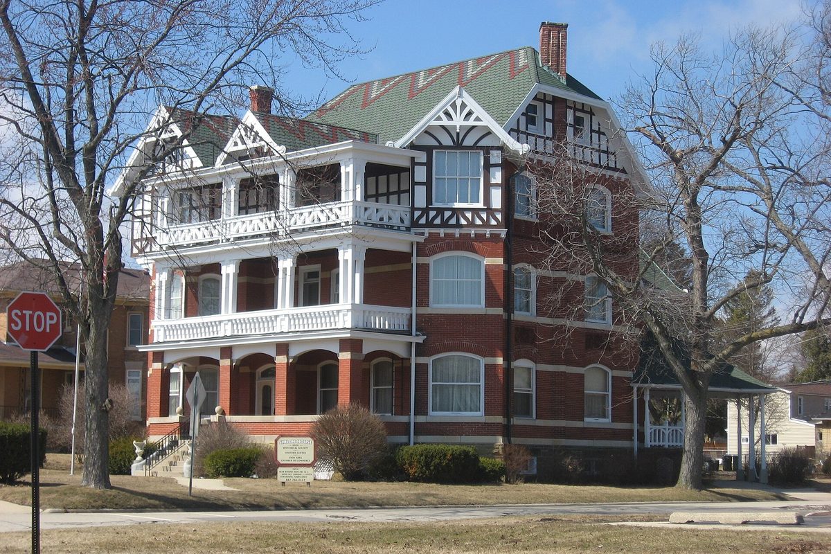 Shiloh House in Zion, a red brick Victorian mansion with white two-story porches and a patterned green roof, beside a stop sign and bare trees