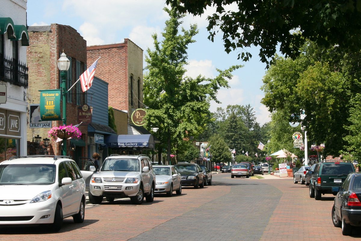 Brick shopfronts, a green Welcome banner and parked cars along Zionsville's brick-paved Main Street on a sunny day